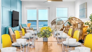 A dining area with a wooden table set for four, yellow chairs, a central plant, and a television on the wall. - Dog-Friendly Beachfront Home with Pool (Gulf Shores)