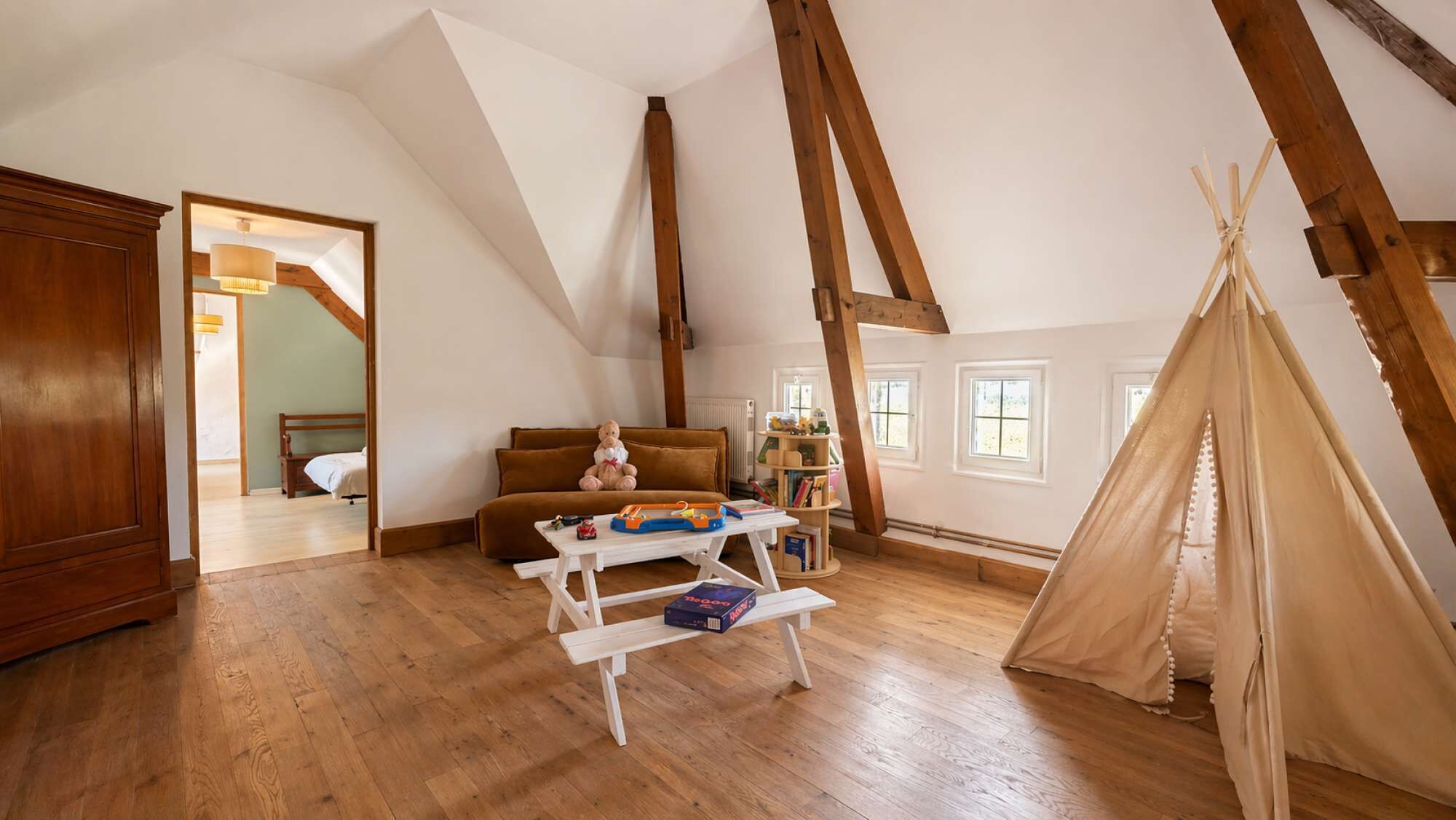 Et loftrum med træbjælker, en sofa, et bord med legetøj, en reol og et tipi.