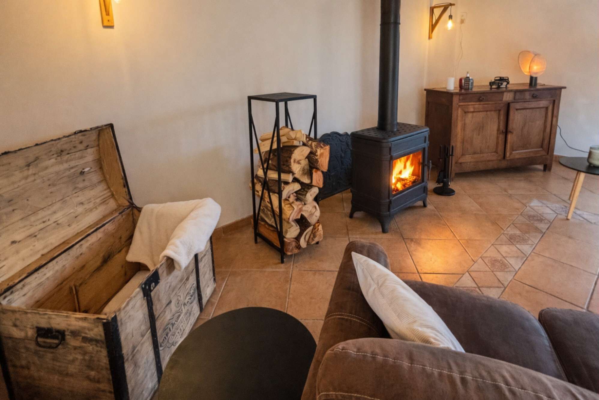 Un salon chaleureux avec un canapé, une malle en bois, une cheminée et une pile de bois de chauffage.