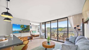 A modern living room with a large TV, a sofa, a dining area, and a balcony with a scenic view. - Waikiki Banyan w/ pool, hot tub & lanai (Honolulu)