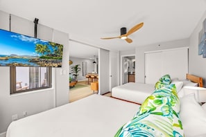Quarto de hotel com uma cama, um ventilador de teto, uma TV na parede e vista para a praia através de uma janela.