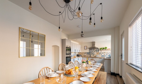 A dining area with a long table set for a meal, a chandelier, and a kitchen in the background.