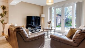 A modern living room with two leather sofas, a flat-screen TV, a small round table, and a large sliding glass door leading to a garden. - Spacious 4-bed, Free Parking, Short or Long Stays (Manchester)