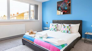 A modern hotel room with a large bed, a view of the city, and a blue accent wall. - Spacious 4-bed, Free Parking, Short or Long Stays (Manchester)