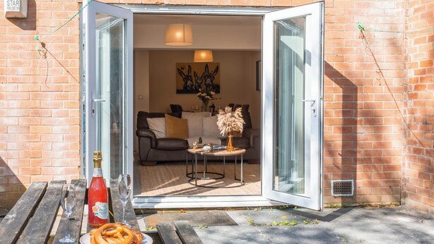 An open glass door leading to a living area with a sofa, a small round table, and a bottle of red wine and glasses on a wooden bench outside. - Spacious 4-bed, Free Parking, Short or Long Stays (Manchester)