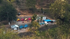 Un área rural con varias construcciones pequeñas, algunas con techos azules y otras con techos rojos, rodeada de vegetación y colinas.