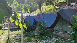 Un camping con varias tiendas de campaña, algunas cubiertas con lonas azules, ubicado en una zona cubierta de hierba, rocas y vegetación.