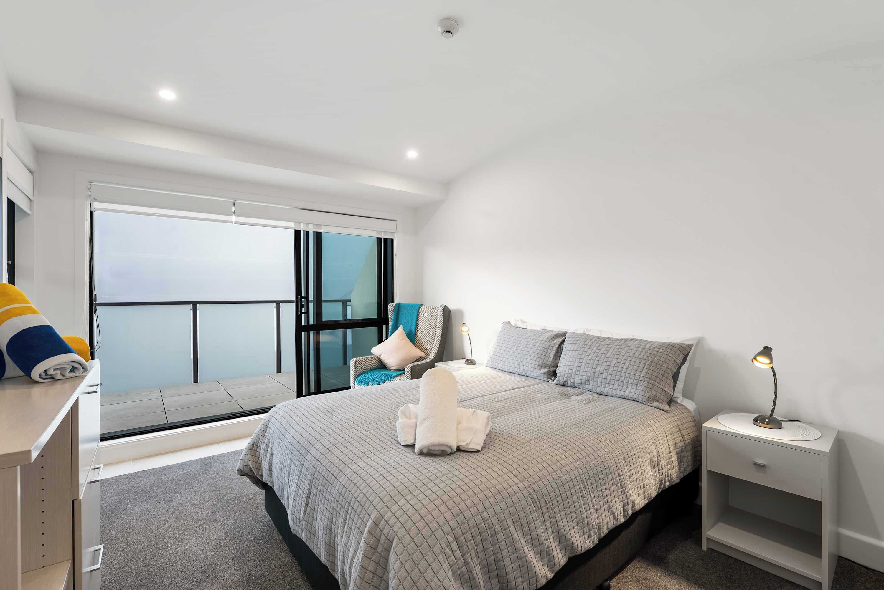 A modern bedroom with a large bed, a bedside table with a lamp, a chair with a view, and a window with blinds.
