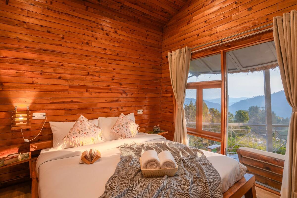 A wooden cabin bedroom with a bed, pillows, a bedside table, a window with a view of the outdoors, and a window blind.