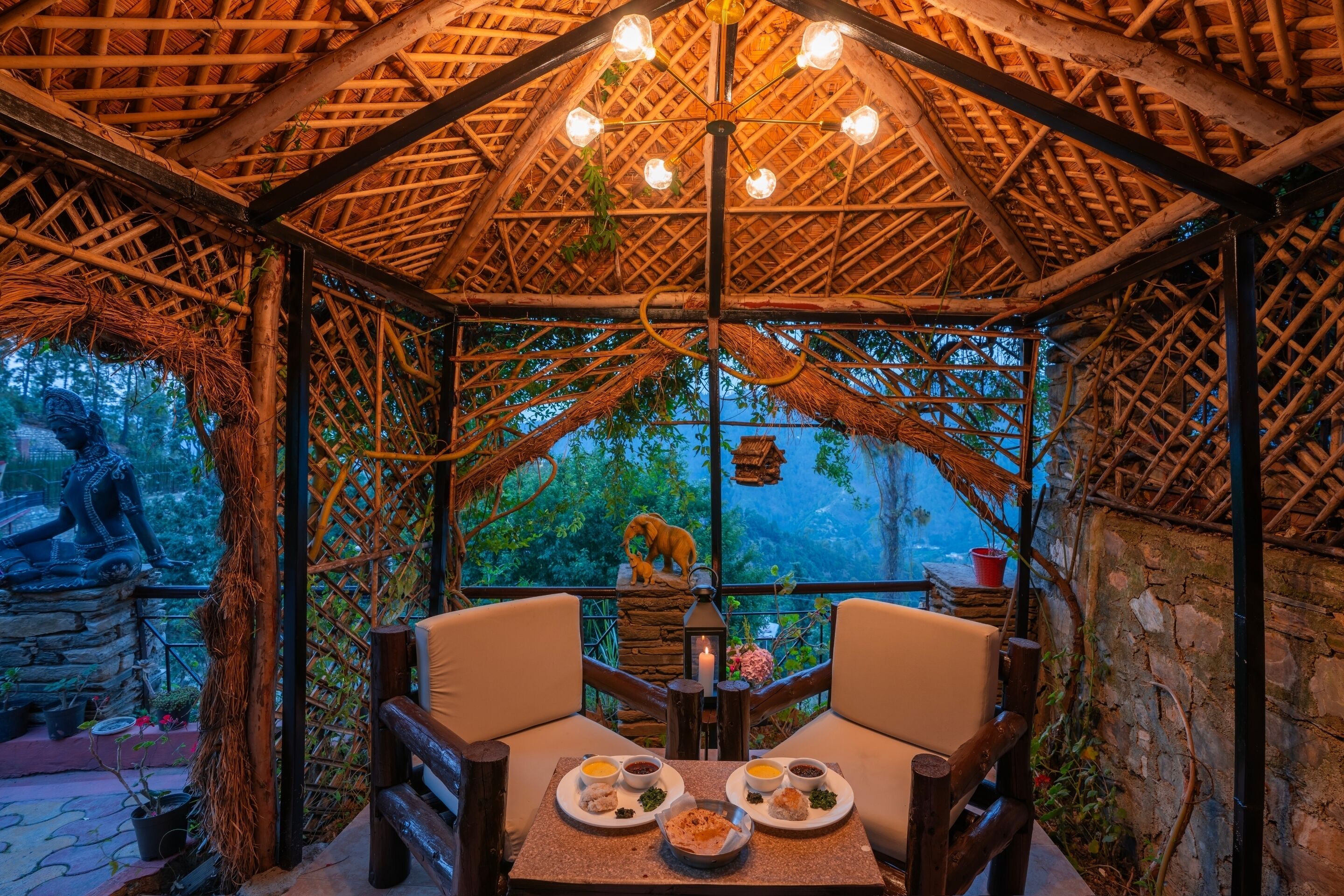 A thatched-roof gazebo with a table set for two, surrounded by stone walls and greenery.