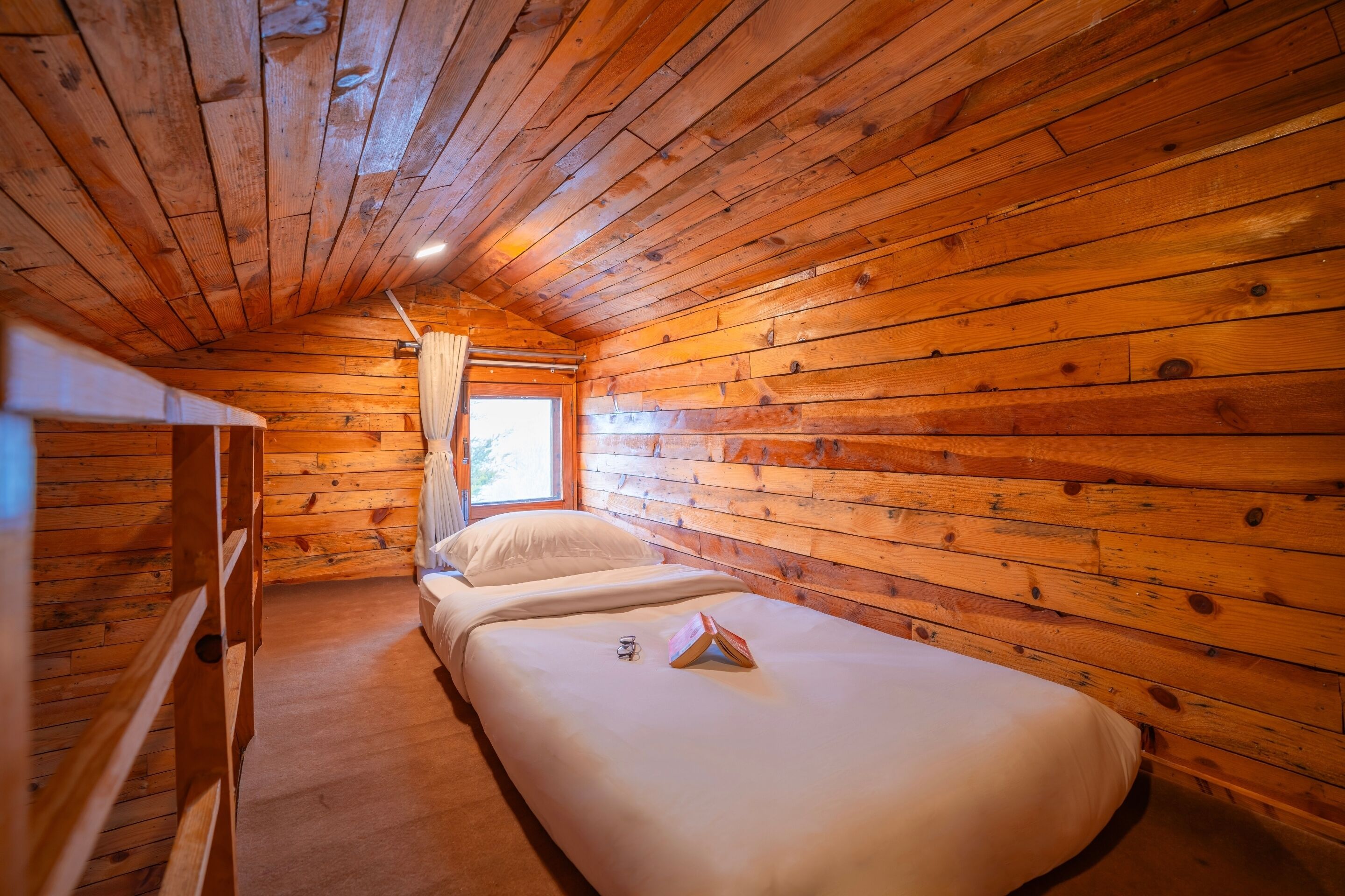 A cabin with wooden walls and a sloped ceiling. There are two beds with white bedding and a small table between them.