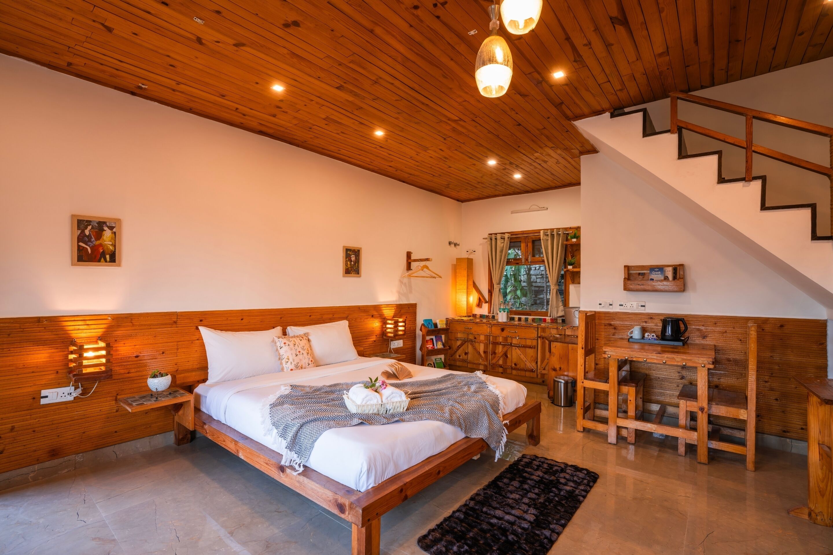 A bedroom with a wooden bed, bedside lamps, a small table, and a staircase.
