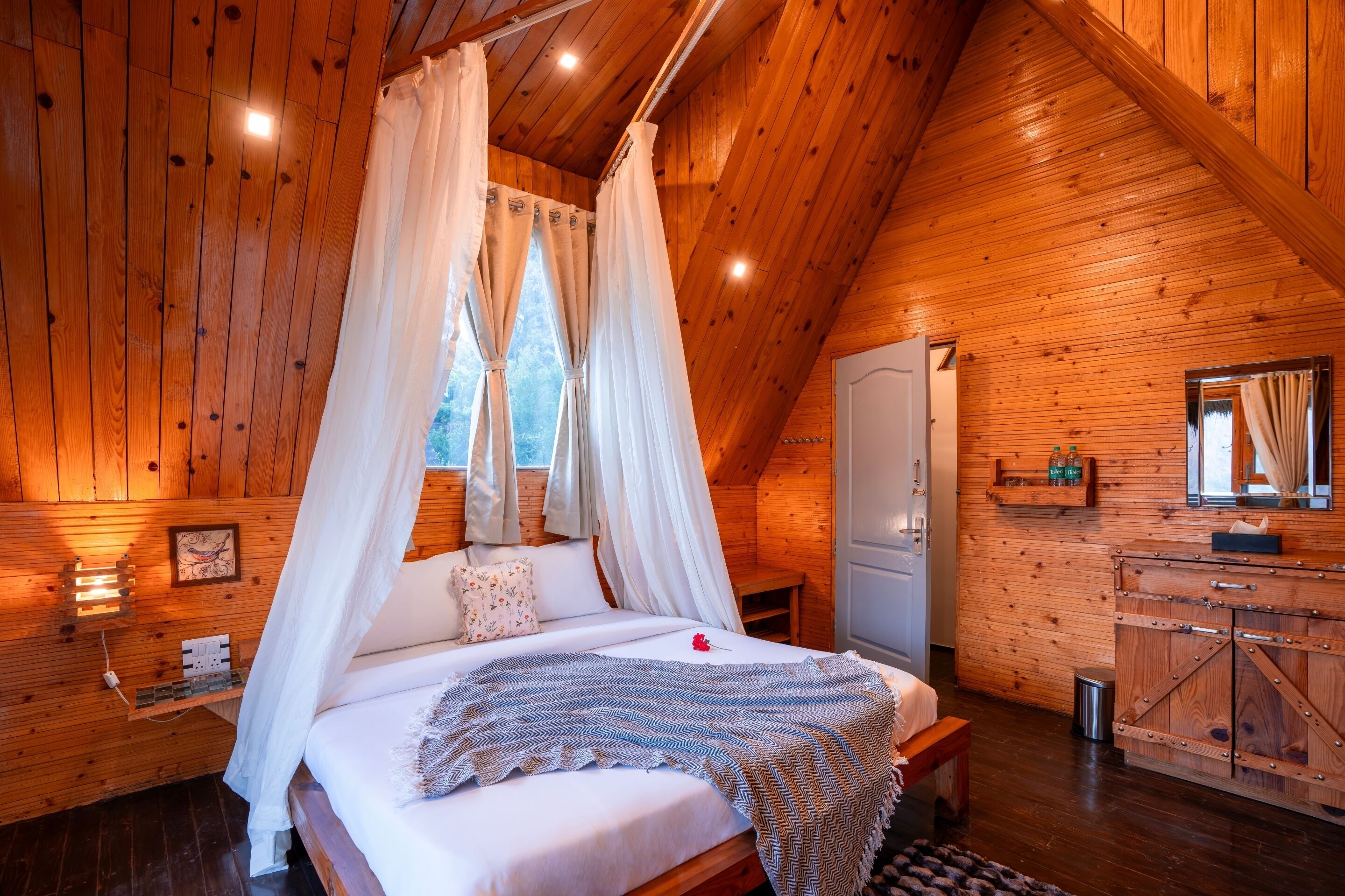A wooden cabin bedroom with a four-poster bed, a bedside table, and a small window with curtains.