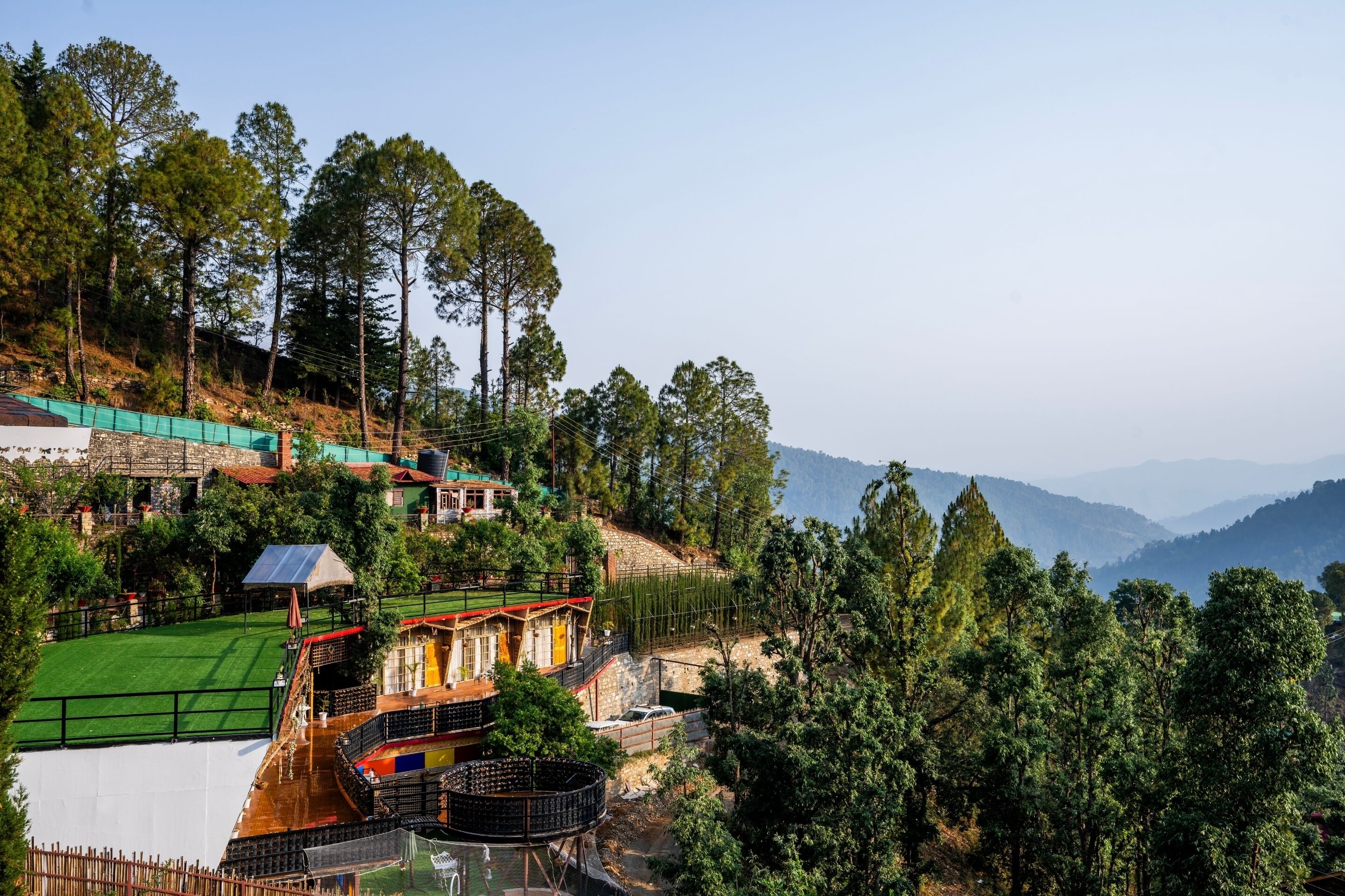 A hillside hotel with a green roof, a swimming pool, and a view of the surrounding forest and mountains.