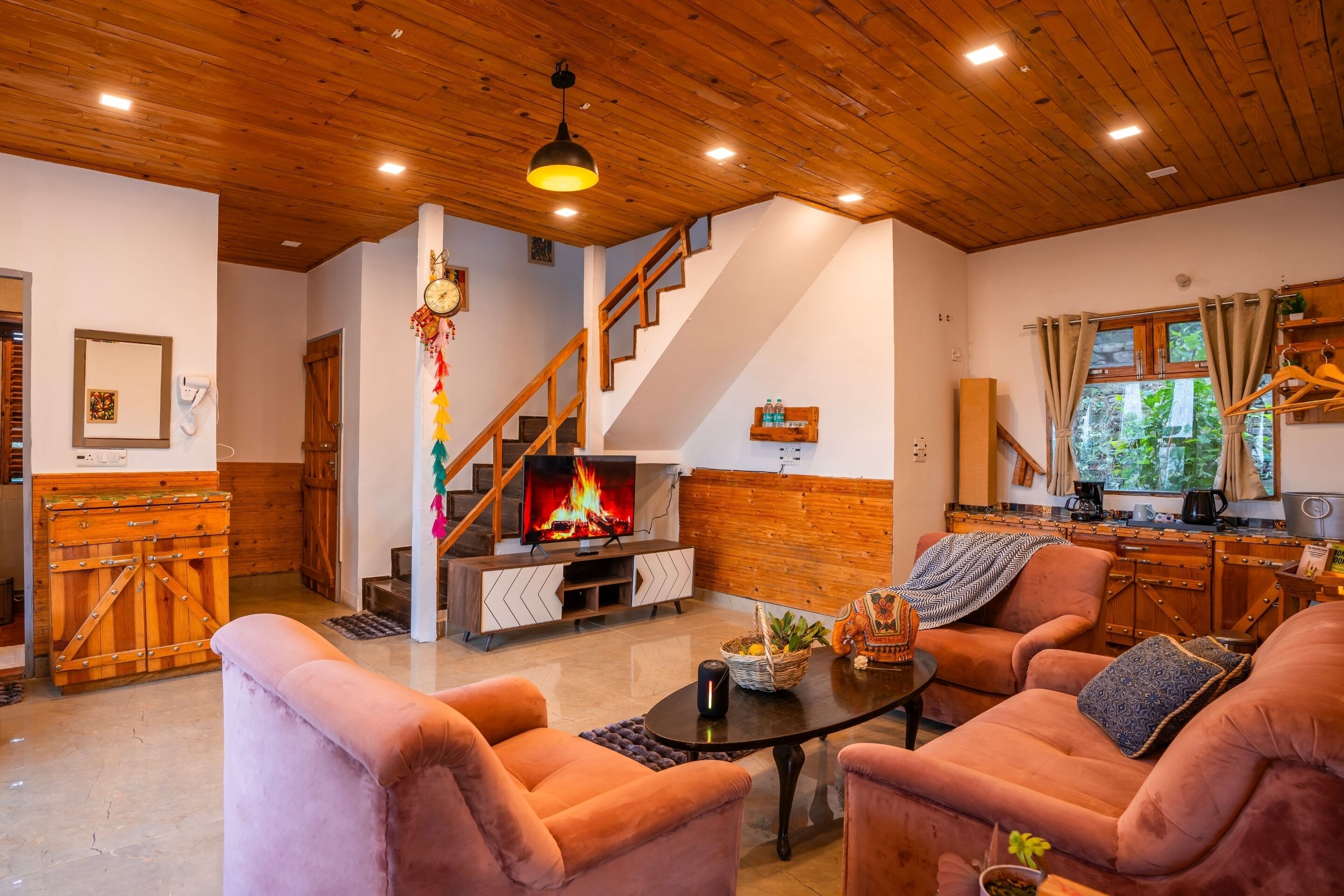 A cozy living room with a fireplace, plush seating, a staircase, and a kitchen area.