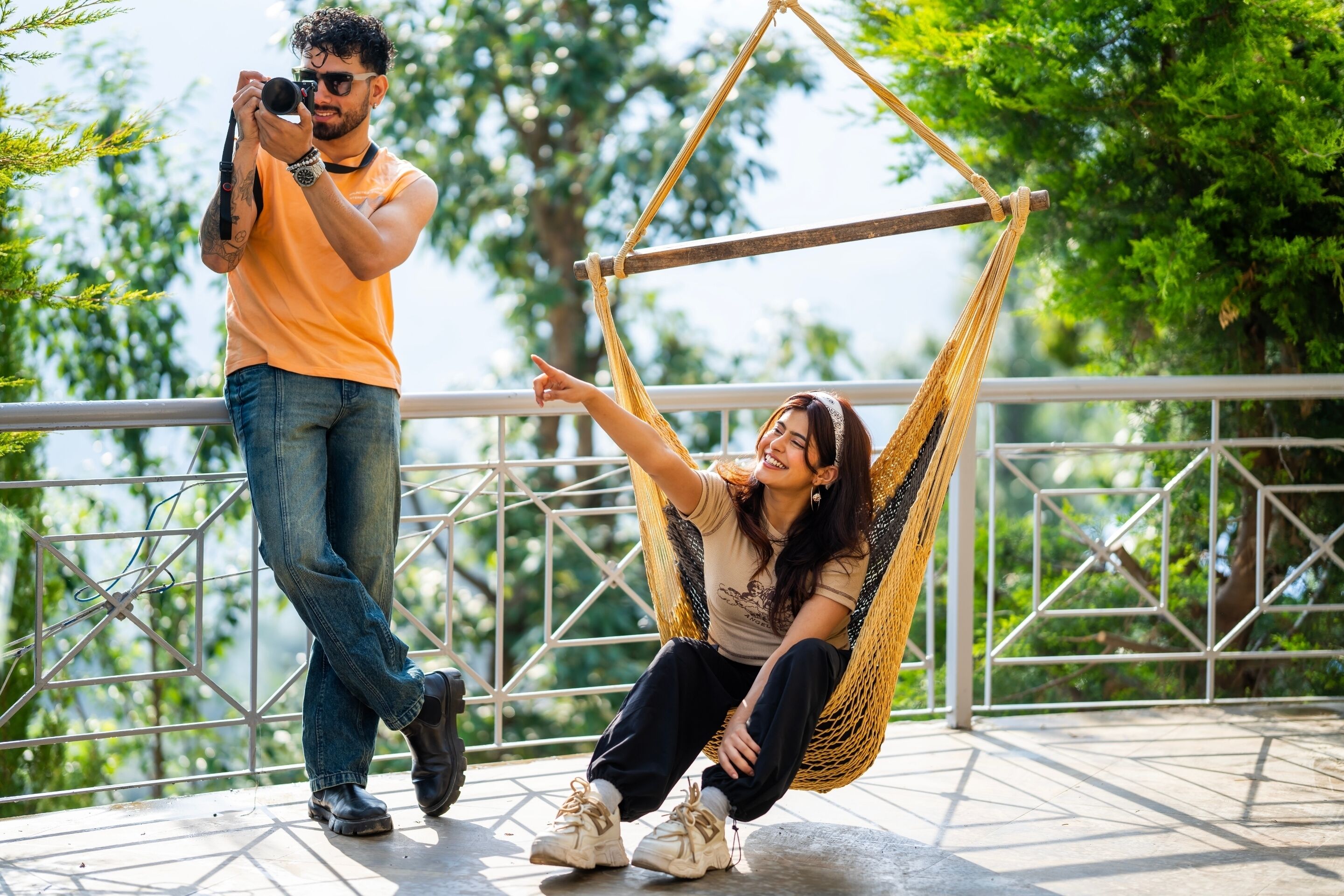 A person in a hammock on a balcony with a railing, another person taking a photo, and greenery in the background.