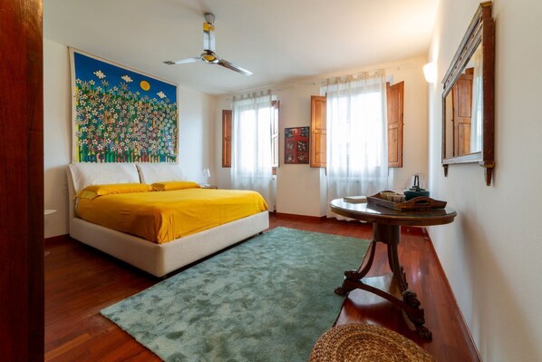 A bedroom with a bed, a ceiling fan, a round table with a lamp, and a painting on the wall.