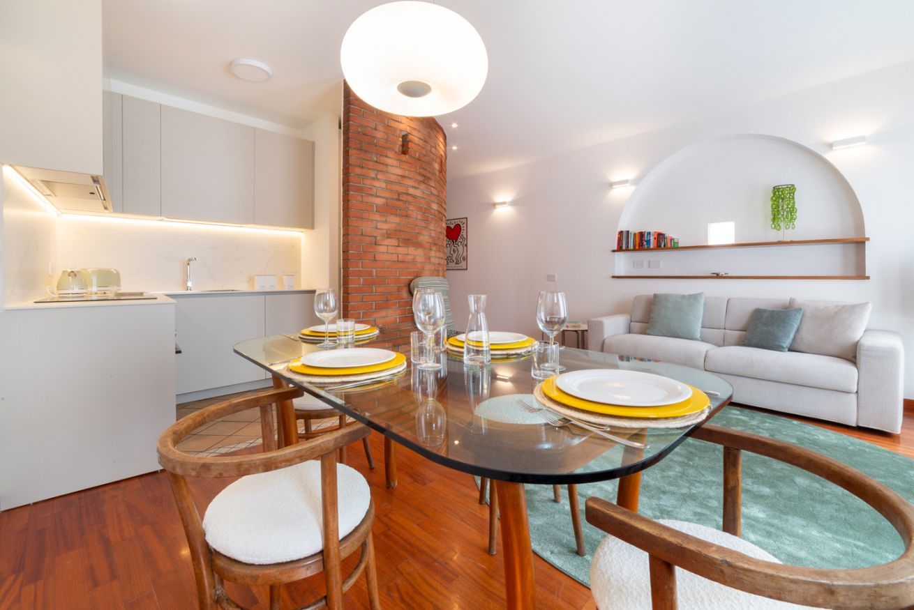 Una cucina moderna con mobili bianchi, una parete decorata con mattoni e un'area pranzo con un tavolo in vetro apparecchiato per quattro persone.