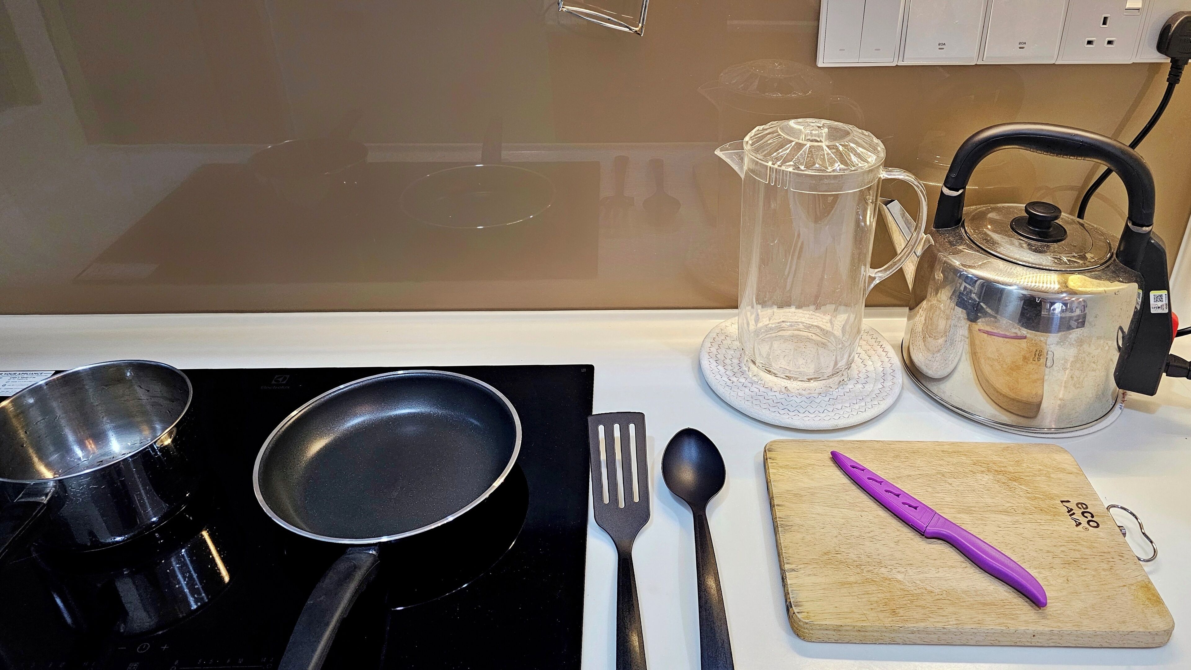 Keittiön työtasolla on paistinpannu, lasikannu, ruostumattomasta teräksestä valmistettu kattila, puinen leikkuulauta, jossa on violetti keittiöväline, ja lusikka.