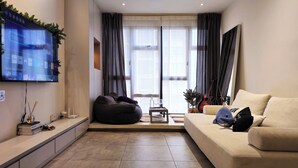 A modern living room with a flat-screen TV, a sofa, a bean bag, a small table with glasses, and a window with curtains.