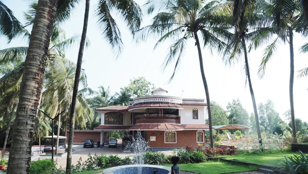 A two-story house with a circular water fountain in the front yard, surrounded by palm trees and a well-maintained garden. - Sugandhi Bhoja Shetty (Mangaluru)