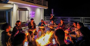 A group of people seated around a fire pit at night, with a neon sign in the background. - goSTOPS Mcleodganj Bhagsu (Dharamshala)