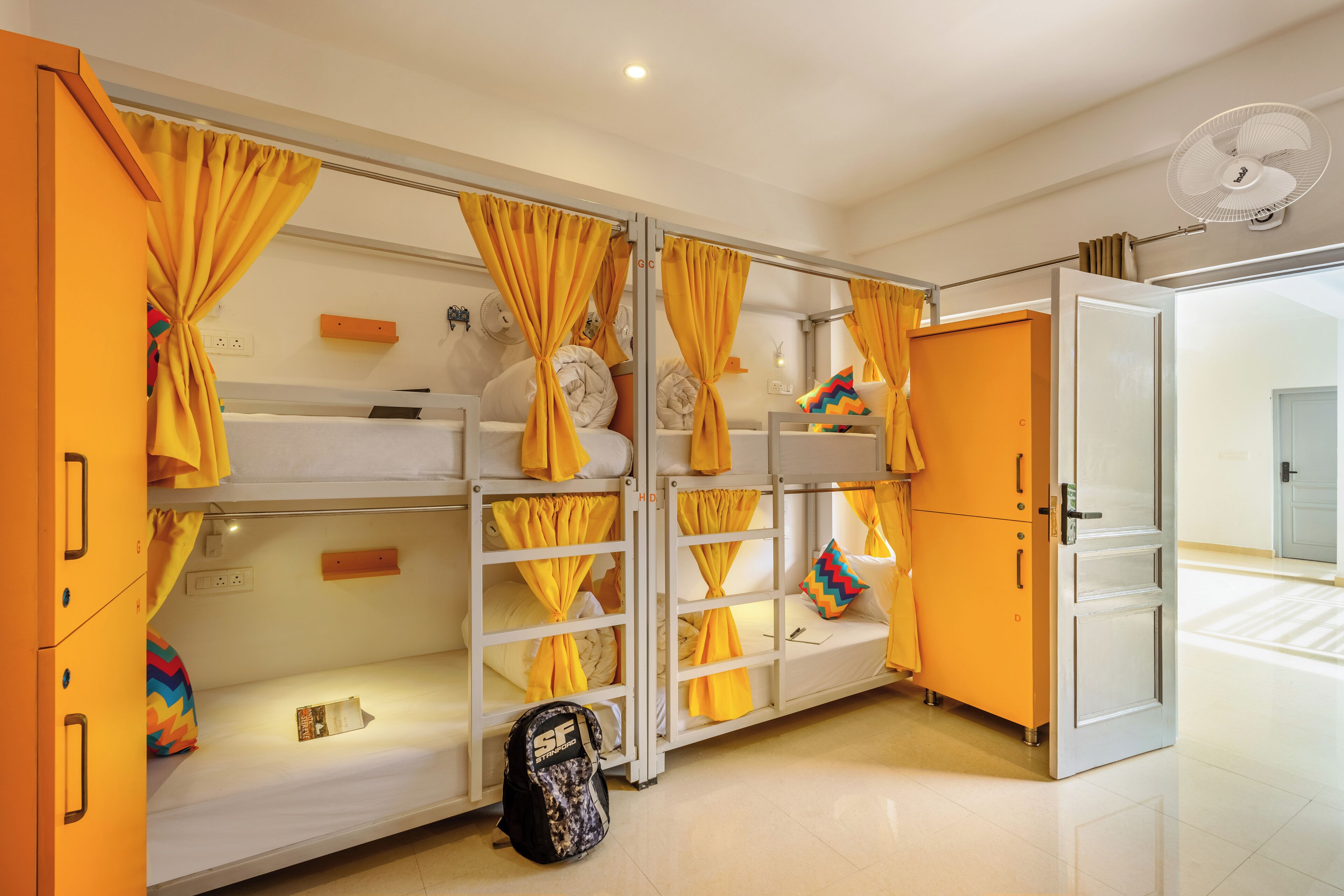 Une chambre avec des lits superposés, des rideaux jaunes, un ventilateur blanc et un sac à dos posé sur le sol.
