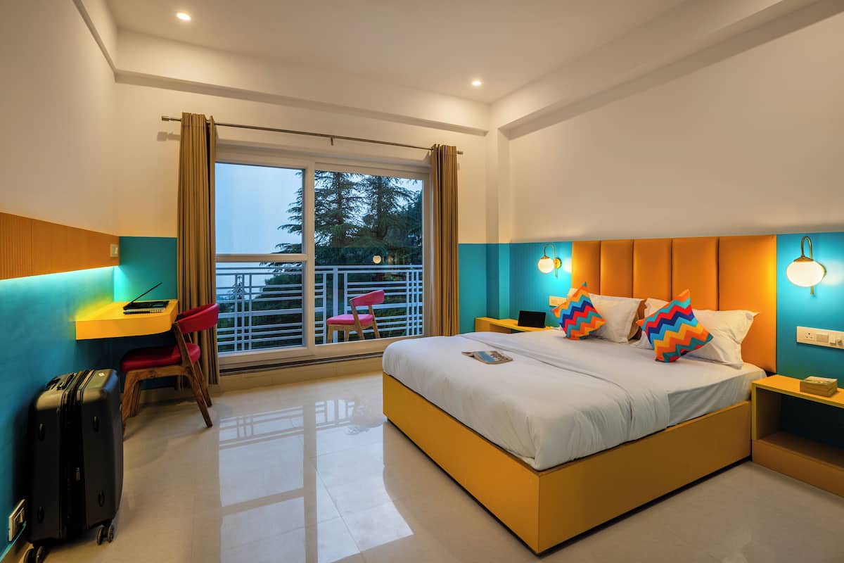 A modern hotel room with a large bed, a desk, a chair, a suitcase, and a balcony with a view of trees.