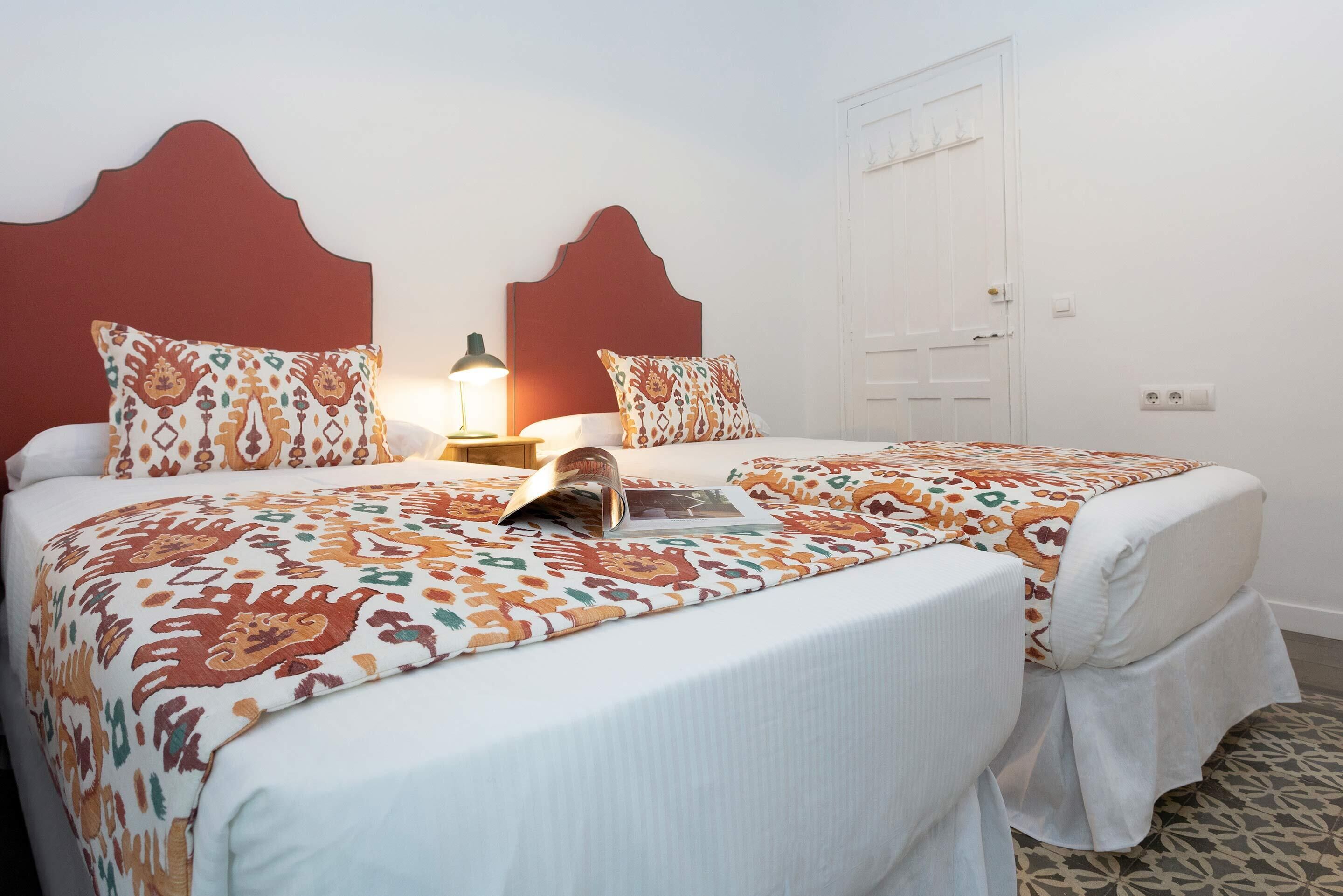 A double room with two beds, each with a patterned bedspread and matching pillows. There is a bedside table with a lamp, and a door to the right.