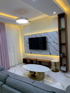 A modern living room with a marble floor, a round coffee table, a sofa, a TV mounted on a marble wall, and a wooden bookshelf.