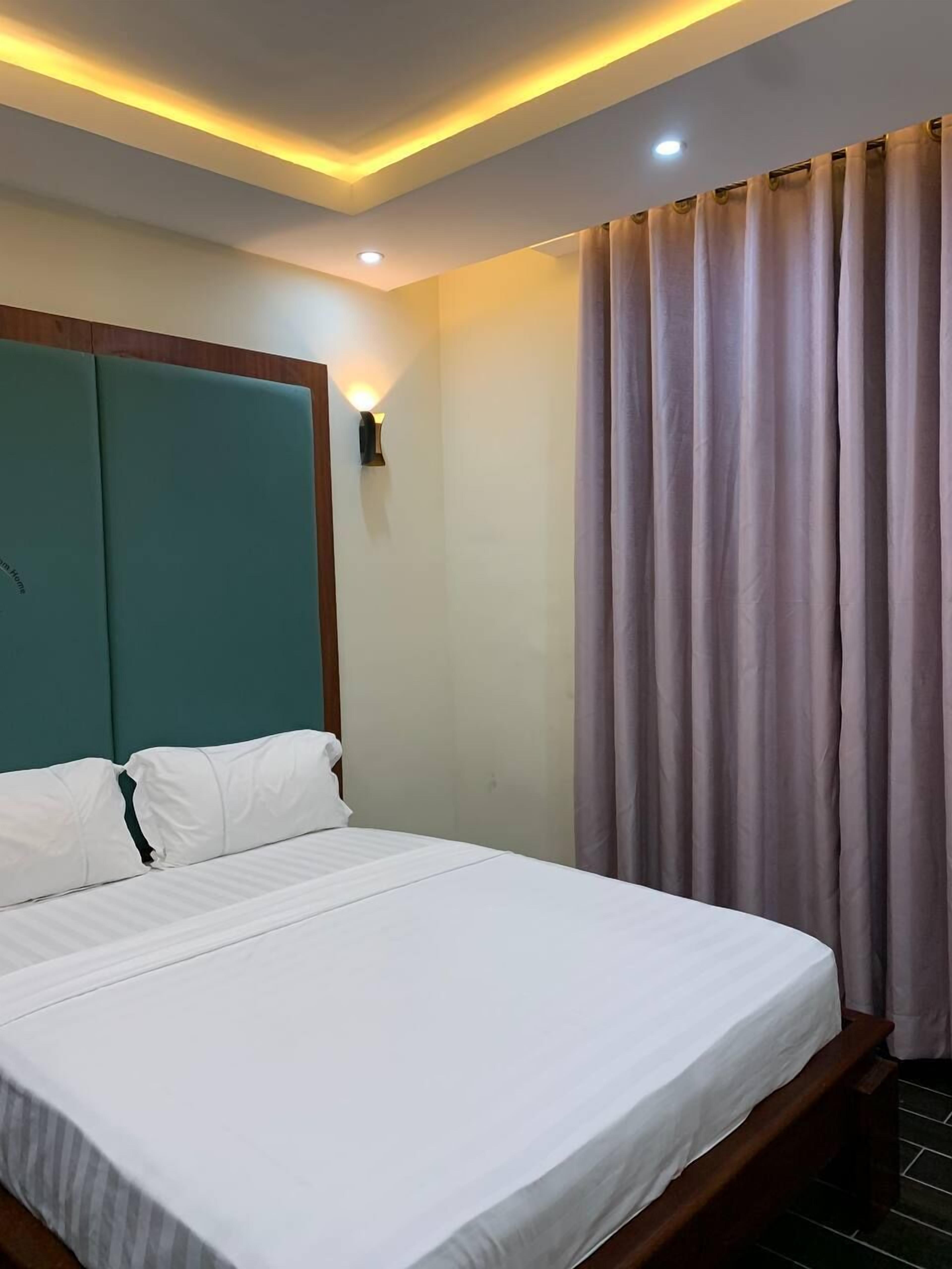 Habitación de hotel con una cama grande, cabecero color turquesa, ropa de cama blanca, una lámpara fijada a la pared y una ventana con cortinas.