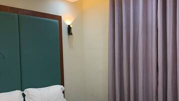 Habitación de hotel con una cama grande, cabecero color turquesa, ropa de cama blanca, una lámpara fijada a la pared y una ventana con cortinas.
