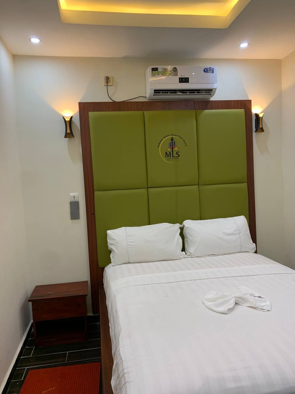 A hotel room with a green padded headboard, white bedding, a white towel on the bed, an air conditioning unit above the headboard, and two wall-mounted lights.