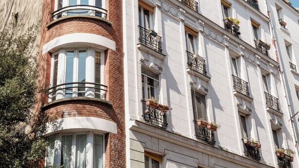 Un immeuble à plusieurs étages dont la partie d’angle présente une façade incurvée, de nombreux balcons dotés de garde-corps noirs et des fenêtres à cadres blancs.