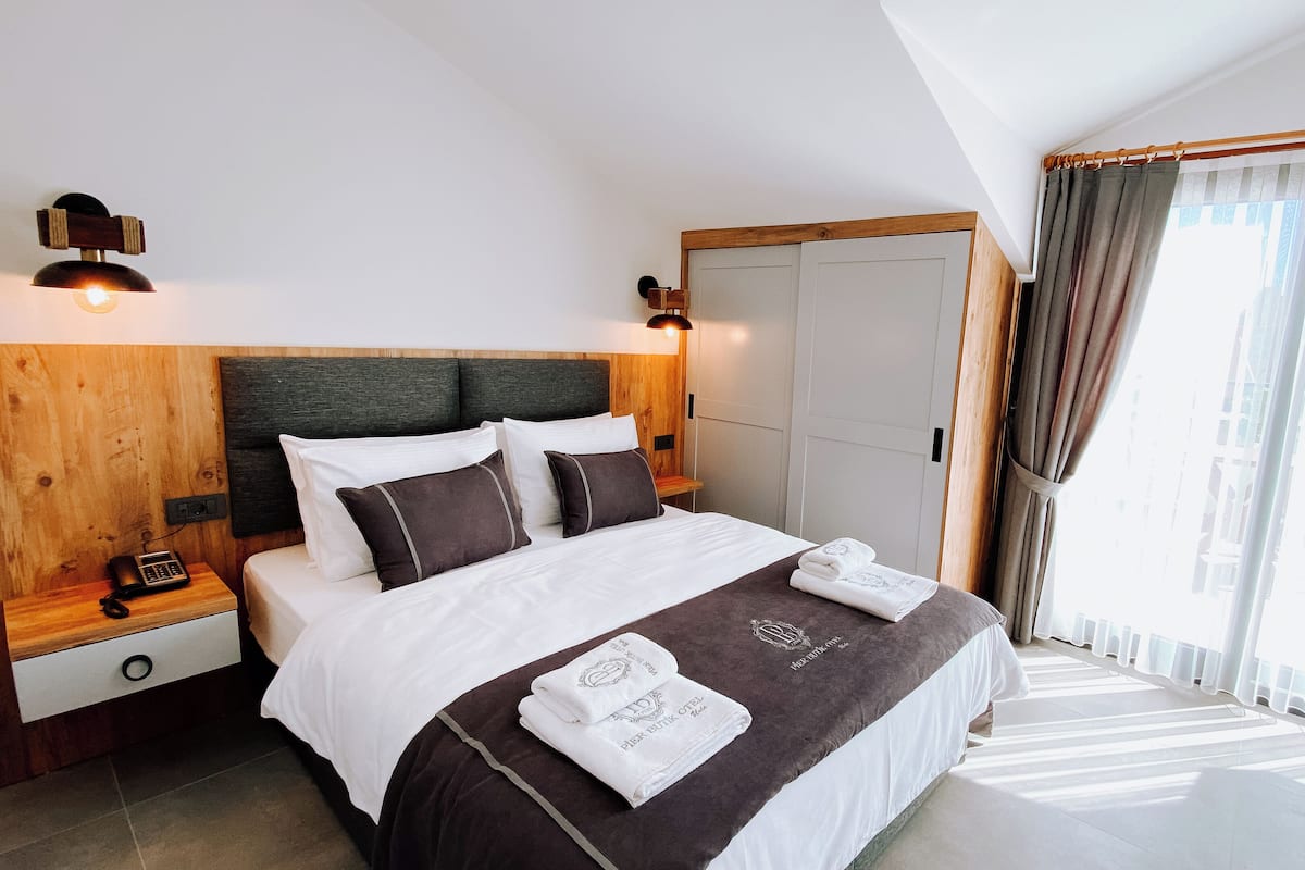 A modern hotel room with a wooden headboard, a bed with white and dark brown bedding, a bedside table with a phone, and a view of the city through large windows.