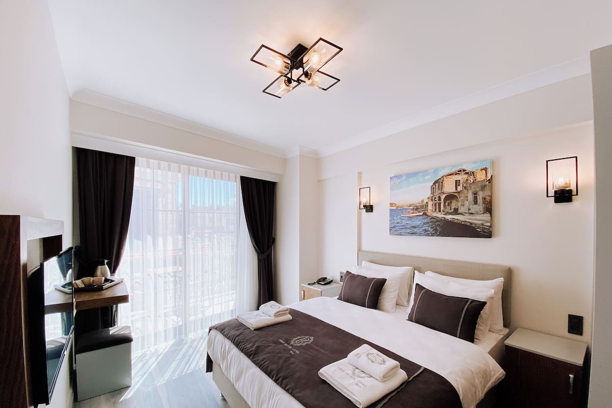 A hotel room with a large bed, a desk with a chair, a window with curtains, a painting on the wall, and a ceiling light fixture.