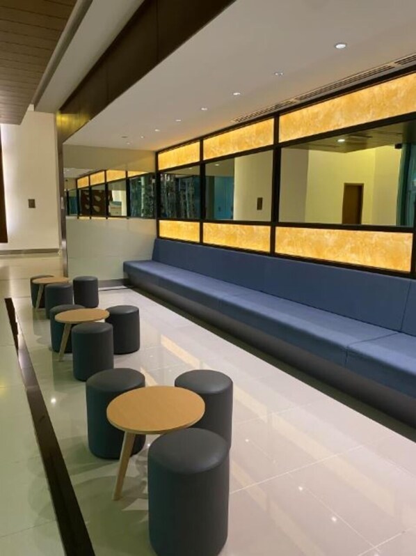 A modern waiting area with a long bench, several round tables, and a glass partition with illuminated panels.