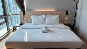 Habitación de hotel con una cama grande, dos almohadas, una toalla enrollada, una mesita de noche, una lámpara y vistas de la ciudad a través de un amplio ventanal.