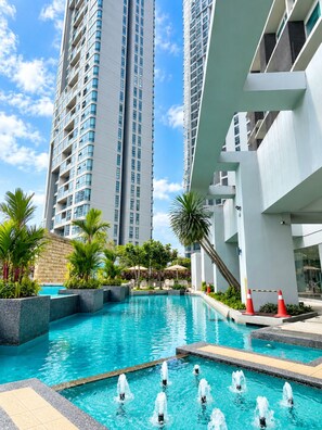 A swimming pool with water fountains, surrounded by palm trees and modern buildings.