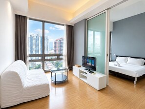 A modern hotel room with a bed, a seating area with a TV, and a large window offering a city view.