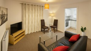 Een moderne woonkamer met een flatscreen televisie, een bank met rode kussens, een glazen salontafel en een eethoek met stoelen en een lamp.