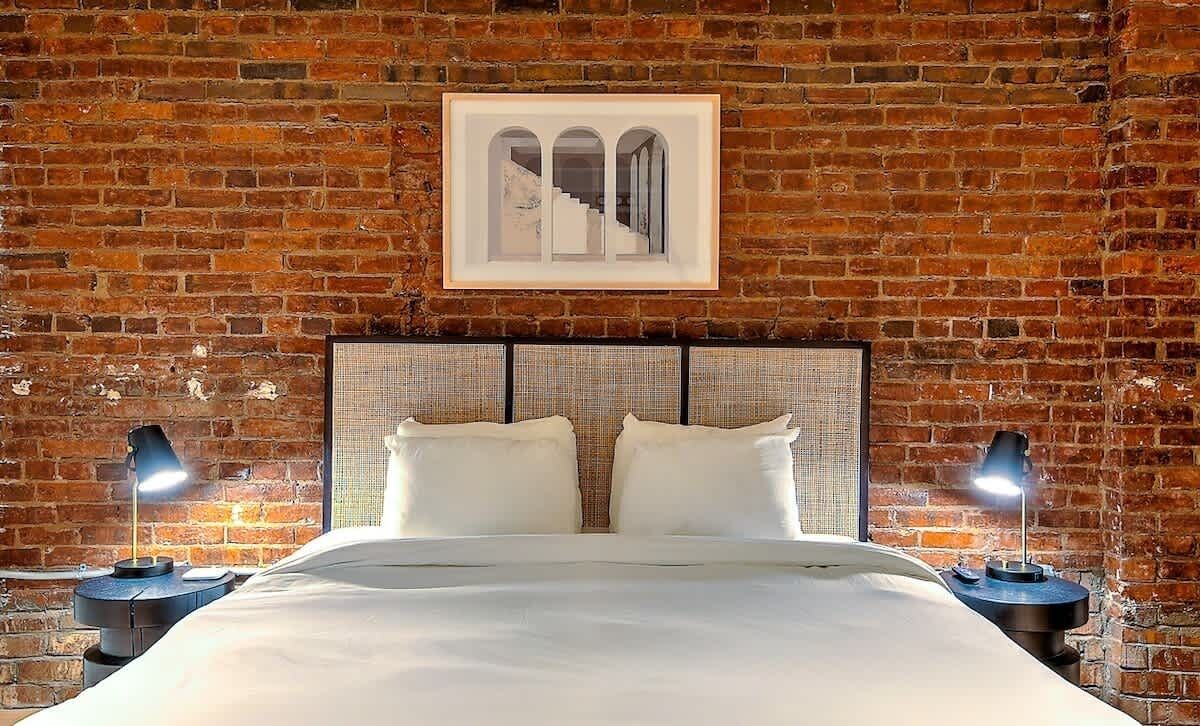 A room with a brick wall, a bed with white bedding, two bedside tables with lamps, and a framed picture above the bed.
