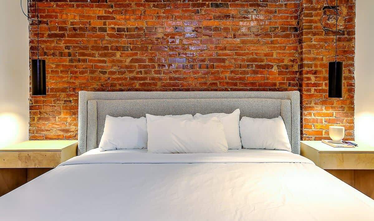 Una cama con cabecera blanca y almohadas, frente a una pared de ladrillo con lámparas colgantes.