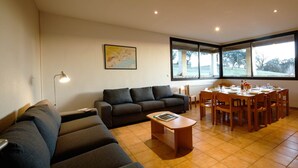 A living room with a sofa, a coffee table, and a dining table set for six. There is a map on the wall and a window providing a view of the outdoors.