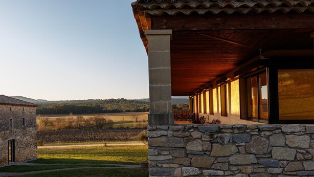 A stone building with a covered patio and a vineyard in the background.