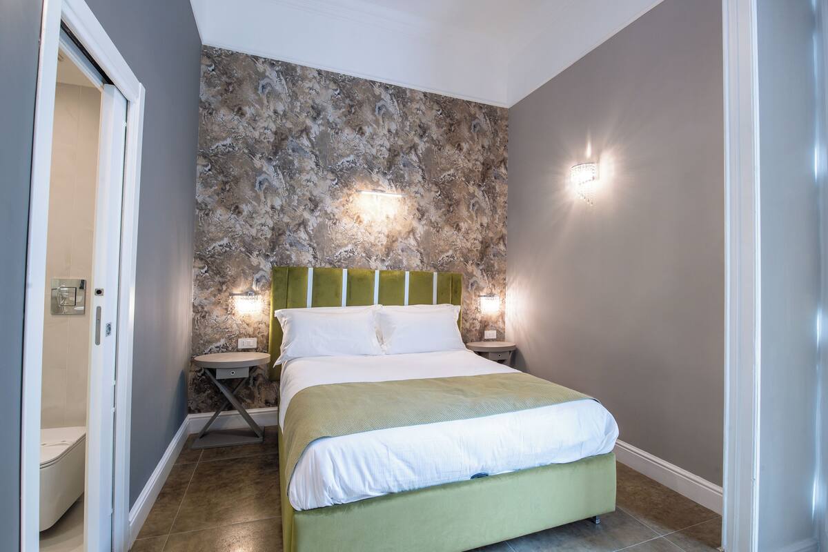 Ein Schlafzimmer mit Bett, Nachttisch, Wandlampe und einer Badezimmertür.