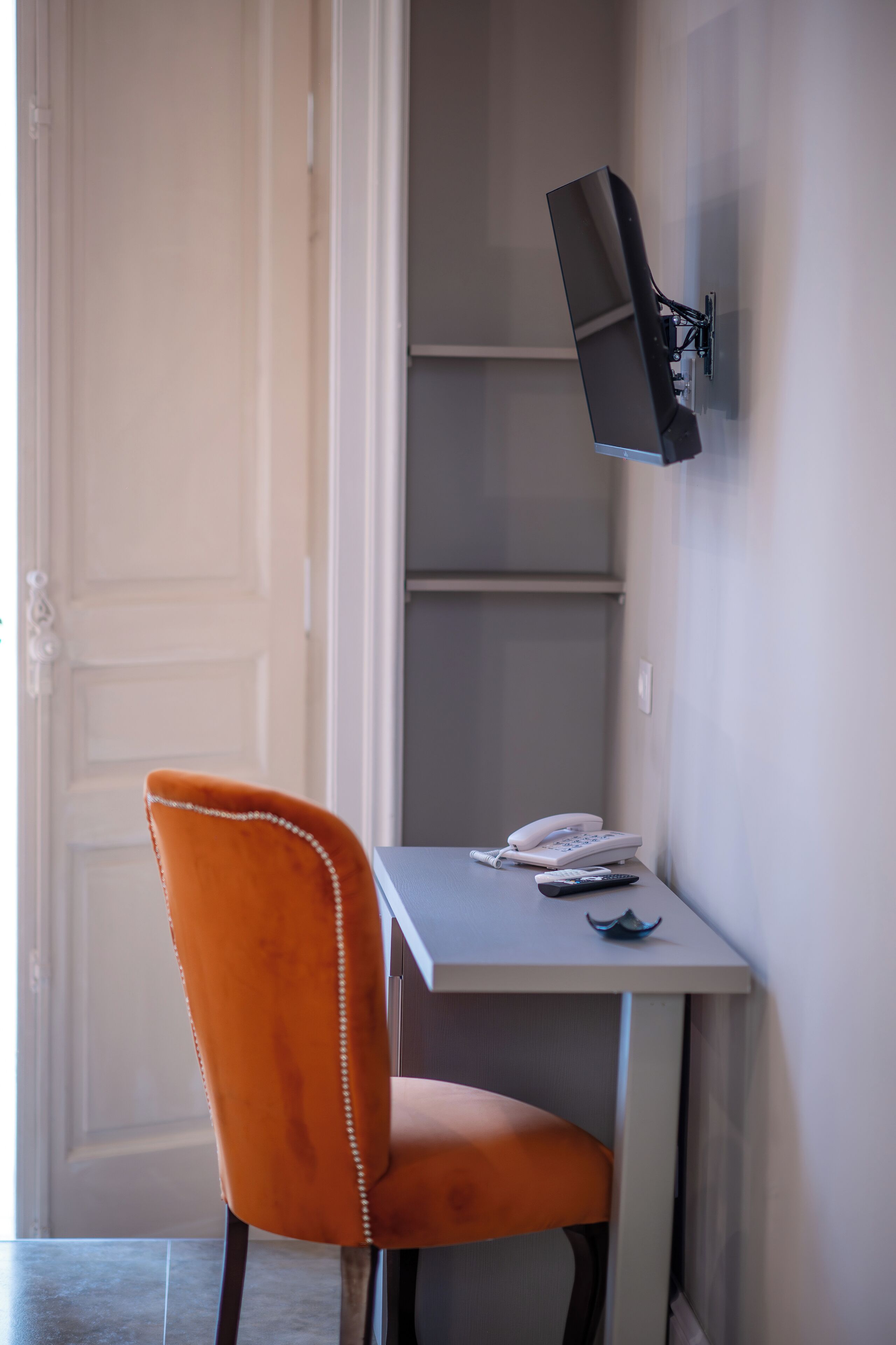 Een kamer met een witte deur, een klein tafeltje met een telefoon en afstandsbediening, en een oranje stoel.