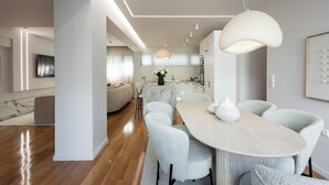 A modern dining area with a wooden table and grey upholstered chairs, a kitchen in the background, and a large pendant light above. - Acropolis Soul (Athens)