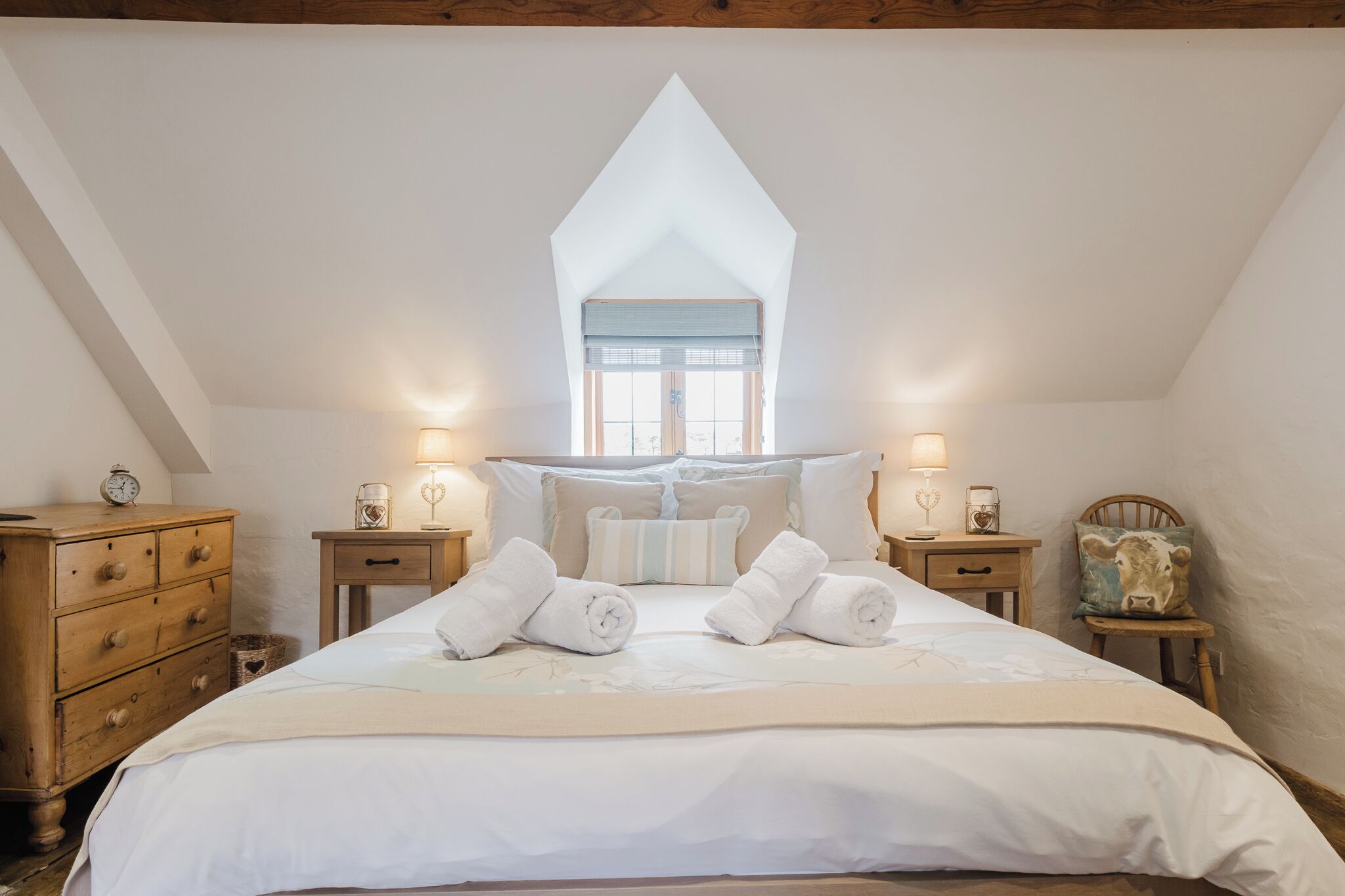 A bedroom with a high, sloped ceiling, a large bed with rolled towels, bedside tables with lamps, and a window with a blue valance.