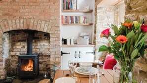 A cozy dining area with a brick fireplace, wooden beams, and a table set for two with plates, cutlery, and a vase of flowers. - Sweetheart Cottage-Cosy 2BD in Cotswolds (Banbury)